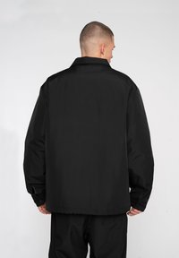 Jachetă neagră, supradimensionată, cu guler, având o textură netedă. Afișată din spate, cu elemente de design minimale și fără logo-uri vizibile.