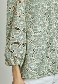 Blusa in pizzo verde con motivi floreali, dotata di dettagli beige chiaro, maniche a tre quarti e chiusura frontale con bottoni.