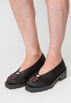 Zwarte gestructureerde lage hak schoenen gedragen op blote voeten met rood-wit kralenontwerp op de tenen tegen een neutrale achtergrond.