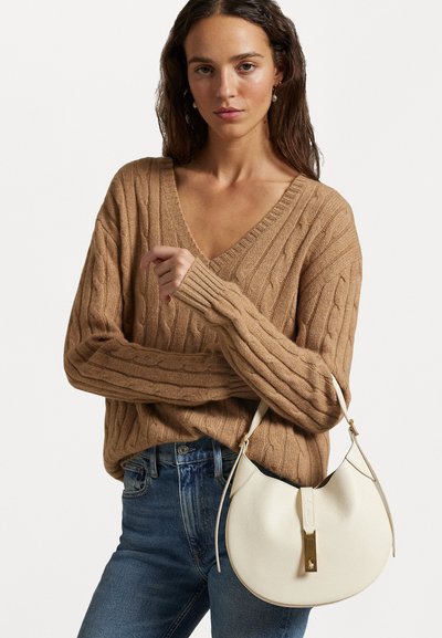 Μπεζ πουλόβερ με πλέξη καλωδίου και V- λαιμόκοψη, συνδυασμένο με καμπύλη λευκή δερμάτινη τσάντα με χρυσές λεπτομέρειες. Ορατά τζην παντελόνια.