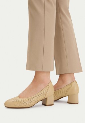 Zapatos beige de tacón bloque con patrón floral perforado, usados con pantalones beige recortados.