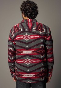 Chemise en polaire rouge et grise à motifs géométriques, avec un col, des poignets à bouton et une texture douce et chaude.