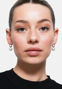 Orecchini a cerchio dorati con pendente di perla, caratterizzati da una superficie texture e una finitura liscia. Il modello indossa un top in maglia nera.