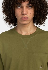 Olivengrøn bomulds t-shirt med rund halsudskæring, korte ærmer, en brystlomme og kontrasterende syninger i et diagonalt mønster.