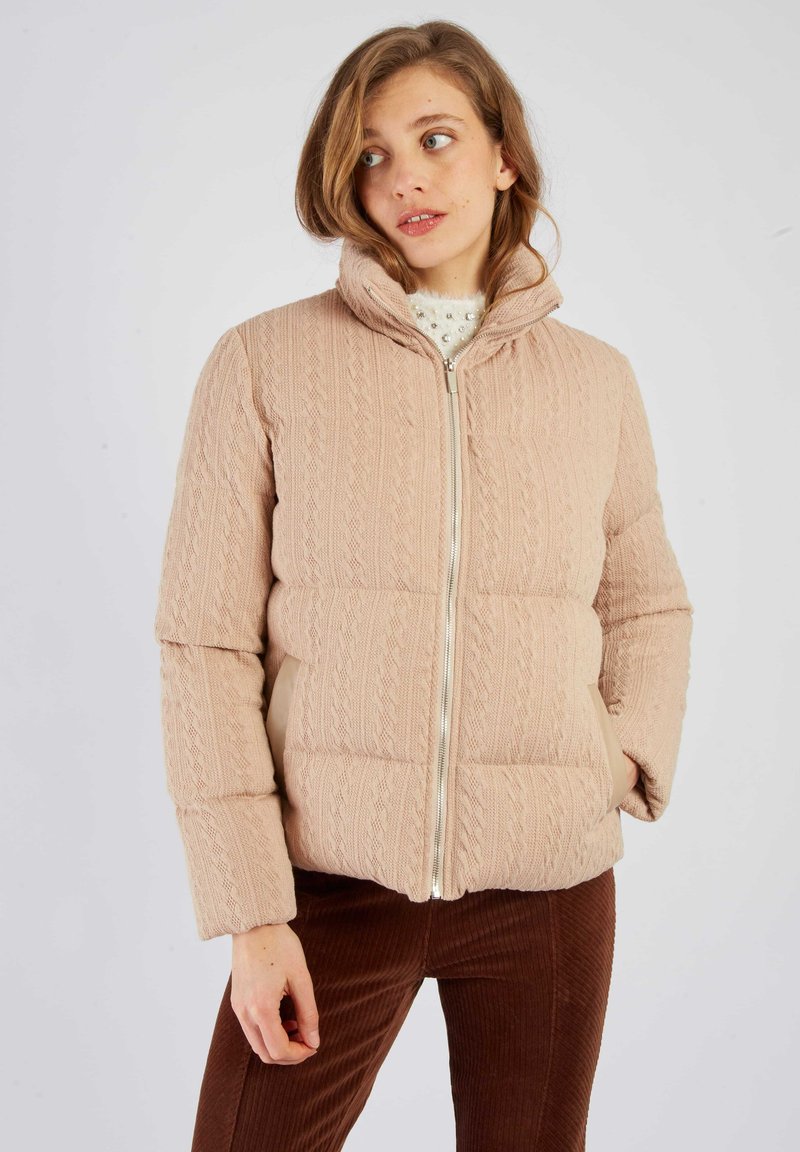Kurtka puchowa z miękkiego różowego dzianiny, z wysokim kołnierzem, zamkiem błyskawicznym i teksturowanym wzorem. Dobana do brązowych spodni sztruksowych.