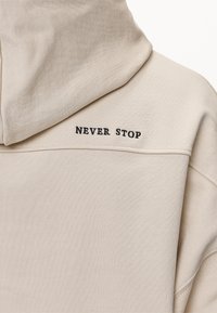 Felpa beige chiaro con un taglio rilassato, texture a coste e la frase "NEVER STOP" ricamata in nero sulla schiena.