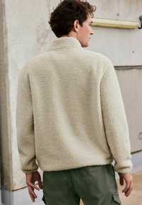 Veste en polaire beige avec un col haut et une surface texturée, dotée d'épaules tombantes et de poignets côtelés, associée à un pantalon vert olive.