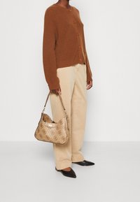 Braune, gerippte Strickjacke mit Knöpfen, kombiniert mit beigen, weit geschnittenen Hosen und einer gemusterten, braunen Handtasche mit goldenen Beschlägen und einem Schulterriemen.