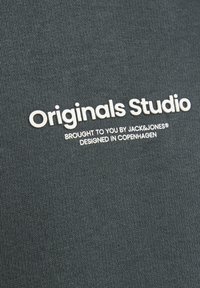 Tessuto grigio con una texture liscia e testo stampato bianco: "Originals Studio" e "Portato a voi da JACK&JONES® Progettato a Copenaghen."