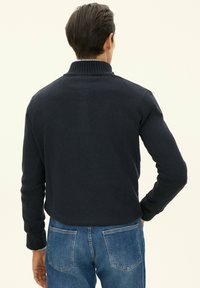 Granatowy sweter z dzianiny z wysokim kołnierzem, długimi rękawami i ściągaczowymi mankietami, noszony z jasnoniebieskimi dżinsami, widziany od tyłu.