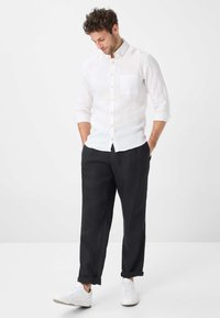 Witte linnen button-up shirt met een borstzak, gecombineerd met zwarte losse broeken en witte sneakers, staande met handen in de zakken.