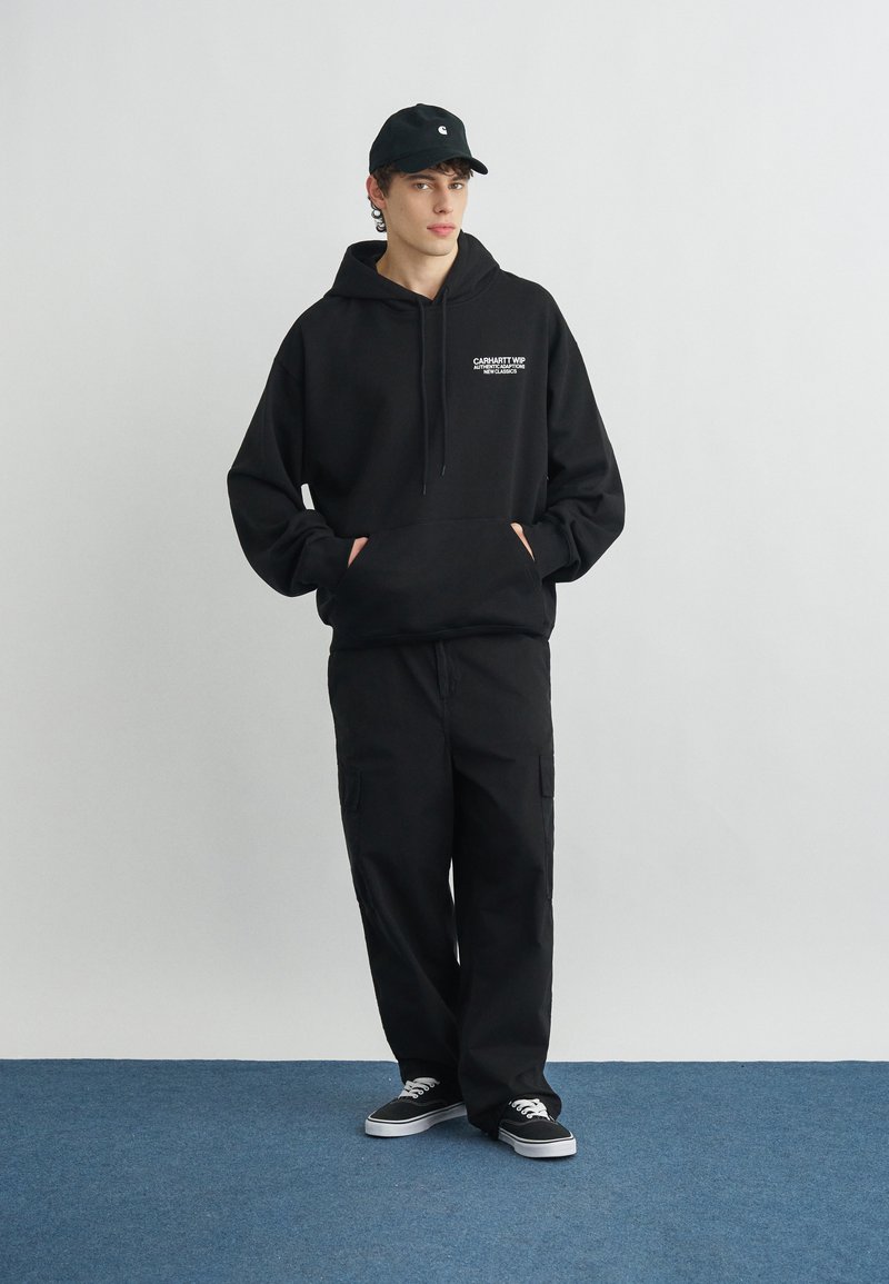 Giovane in piedi con le mani nelle tasche che indossa una felpa nera Carhartt con cappuccio, pantaloni cargo neri, cappellino nero e sneakers nere con lacci bianchi.