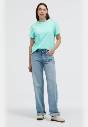 Frau trägt ein hellblaues T-Shirt, das leicht in hoch taillierte, weit geschnittene hellblaue Jeans mit umgeschlagenen Säumen gesteckt ist, und weiße Sneaker.