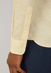 Camicia a righe gialle e bianche con una texture liscia e polsini rotondi, dotata di due bottoni su ciascun maniche.