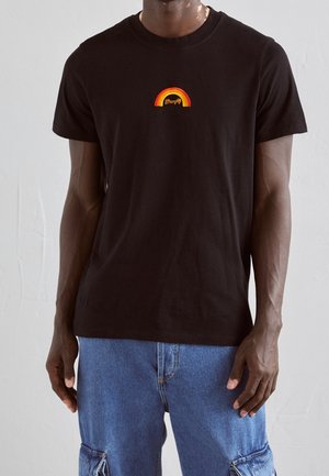 Hombre con camiseta negra con pequeño logo de arcoíris naranja y amarillo y vaqueros cargo azules de pie contra un fondo liso.