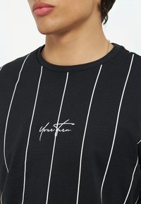 Κοντινό πλάνο ατόμου με μαύρο πουκάμισο με λευκές κάθετες ρίγες και καλλιγραφικό κείμενο "Your Turn" στο κέντρο.