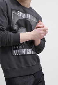 Felpa grigia con stampa grafica che riporta il testo "SPIN IT !" e "ALL NIGHT". Polsini e orlo a costine, materiale misto cotone, vestibilità casual.