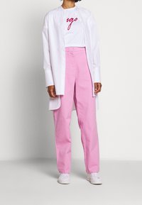 Hvid button-up skjorte lagd over en hvid t-shirt med rød tekst, parret med lyserøde højtaljede bukser og hvide sneakers.