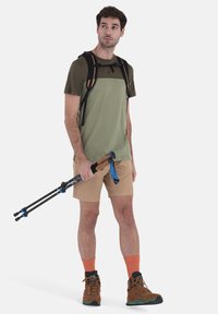 Abbigliamento da escursionismo con una maglietta verde e beige a maniche corte, pantaloni cargo khaki, calze arancioni e stivaletti marroni, mentre tiene in mano dei bastoni da trekking.