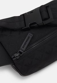 Černá ledvinka s texturovanou látkou, vybavená černým zipem, reliéfním logem a nastavitelným popruhem s plastovou přezkou.