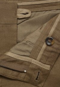 Un tissu marron au fini texturé dispose d'un bouton, d'une poche zippée et d'un détail en boucle ; conçu avec une mise en page simple et pratique.