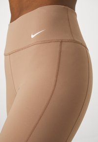 Gros plan d'une personne portant un legging beige taille haute Nike avec des coutures visibles et le logo blanc Nike sur la ceinture.