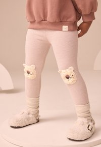 Dieťa nosí pruhované legíny s nášivkami medvieďaťa na kolenách, krémové ponožky, nadýchané krémové papuče a prachovo ružový sveter.