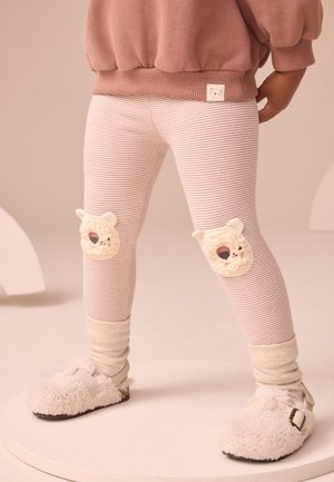 Kind trägt gestreifte Leggings mit Bärengesicht-Patches auf den Knien, cremefarbene Socken, flauschige cremefarbene Hausschuhe und einen staubig rosa Pullover.