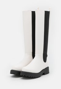 Botas até ao joelho com um cabedal sintético branco, painéis laterais elásticos em preto e uma sola de borracha texturizada em preto. Acentuadas com alças de puxar.