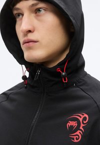 Μαύρο μπουφάν με κουκούλα και κόκκινα κορδόνια, εμπρόσθιο φερμουάρ και κόκκινο tribal σχέδιο στην αριστερή περιοχή του στήθους. Με λεία υφή.