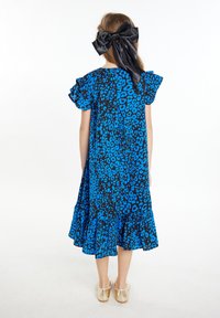 Abito con stampa leopardata blu e nera, con maniche arricciate e orlo merlettato, abbinato a scarpe dorate lucide e a un grande fiocco nero nei capelli.