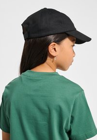 Svart basebollkeps med en böjd skärm och justerbart band, bärs över en grön bomull T-shirt med mjuk textur och rund halsringning.