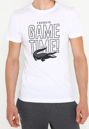Hvid t-shirt med sort tekst "Lacoste GAME TIME!" og en sort krokodillegrafik båret af en person med grå bukser.