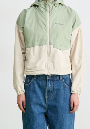 Hættejakke fra Columbia med lysegrøn øvre del og beige nederste del, kombineret med blå denimjeans på en person, der står mod en ensfarvet baggrund.
