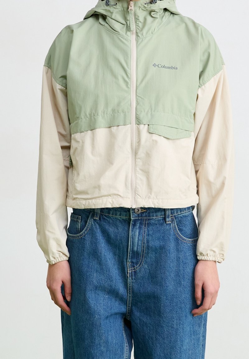 Veste à capuche Columbia avec partie supérieure vert clair et partie inférieure beige, associée à un jean bleu, portée par une personne debout devant un fond uni.
