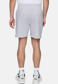 Persona vestida con pantalones cortos deportivos gris claro, calcetines blancos hasta la altura del tobillo con logo y sneakers blancos de FILA, de pie con la espalda hacia el espectador.