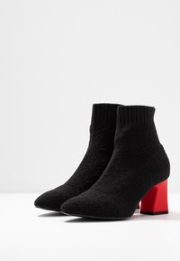 Botas negras hasta el tobillo con un upper de tejido texturizado, cuello acanalado, punta afilada y tacón en bloque rojo a contraste. Suela lisa y forma estructurada.