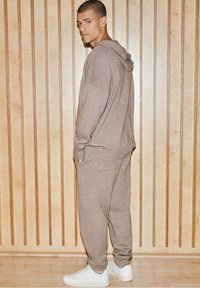 Brązowa, oversize'owa bluza z kapturem oraz pasujące dresy wykonane z miękkiego materiału; zestawione z białymi sneakerami. Tło z jasnych paneli drewnianych.