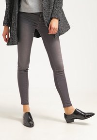 Vaqueros ajustados grises con textura sutil, combinados con zapatos negros pulidos. El conjunto se complementa con un abrigo oscuro con patrón y una camisa gris.