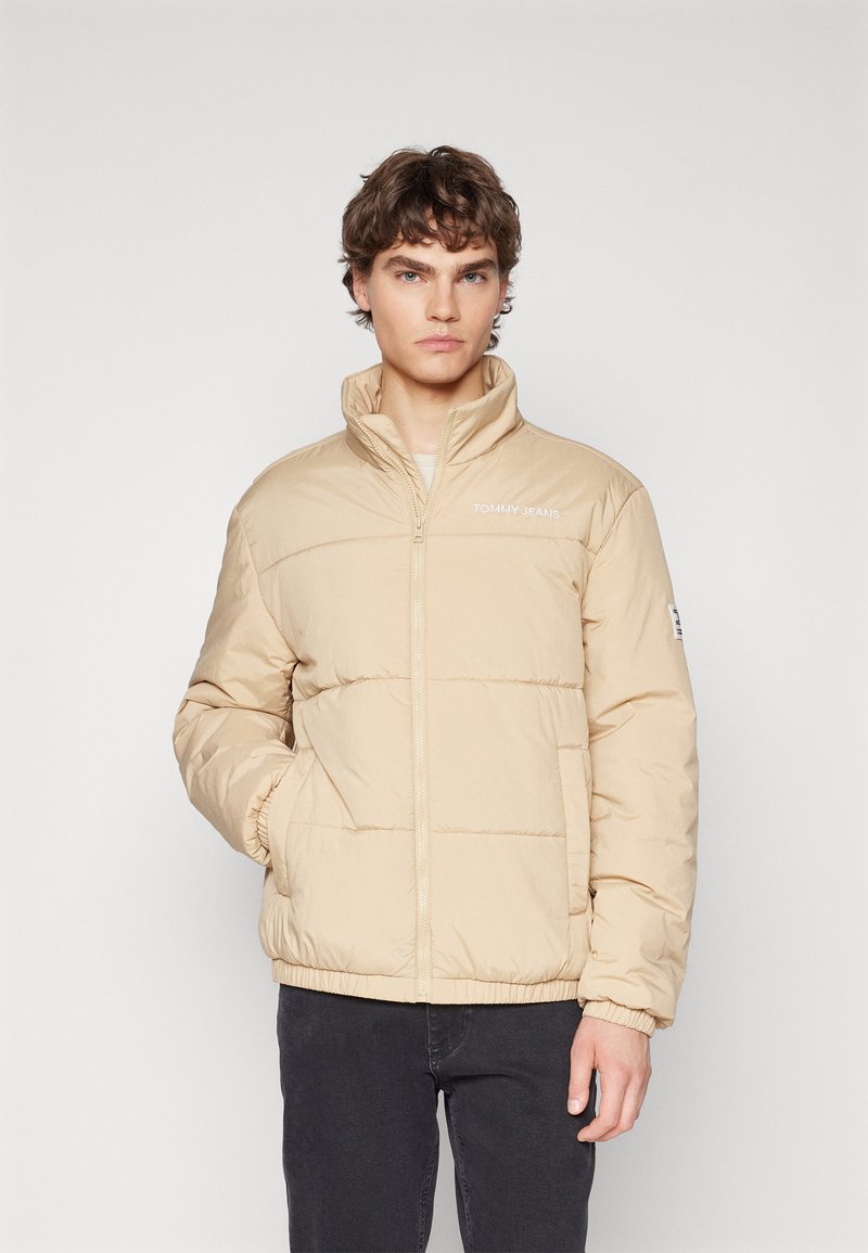 Beige padded jacket with high collar, zip closure, and elastic cuffs. Features logo on chest and a subtle shiny texture.