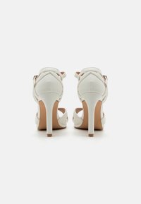 Hvite høyhelte sandaler med kryssede stropper, glatt skinnfinish, spiss tå og slank stiletthæl. Myk beige innersåle.
