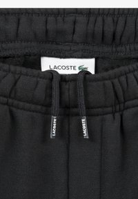 Čierne teplákové nohavice vyrobené z mäkkej látky, so elastickým pásom, nastaviteľnými šnúrkami a štítkom Lacoste so zeleným krokodílom.