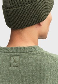 Berretto lavorato a maglia verde oliva con una texture a coste, abbinato a un maglione coordinato con un piccolo emblema sulla schiena.