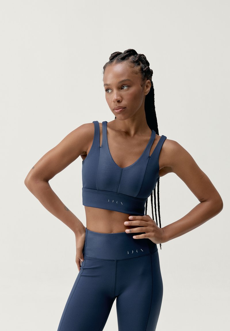 Tamsiai mėlynas sportinis liemenukas su plačiomis petnešomis ir išlenktu iškirpimu, derinamas su suderintomis aukšto juosmens kelnaitėmis. Lygi, elastinga medžiaga.