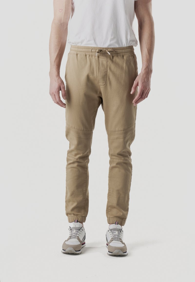 Pantalones jogger beige con cintura elástica, piernas cónicas y costuras laterales; usados con zapatillas grises y blancas con detalles en rojo.