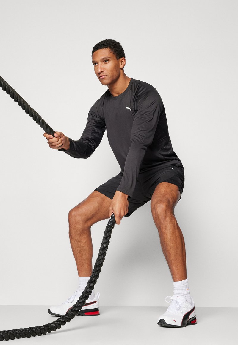 Czarna bluza z długim rękawem, czarne szorty, trzymając grube czarne liny treningowe. Czerwono-białe buty sportowe. Krótkie brązowe włosy, zaangażowany w ćwiczenia.