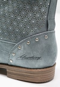 Botín de cuero gris hasta el tobillo con diseño floral perforado, acentos plateados y un tacón de madera. Presenta tachuelas decorativas y una firma grabada.