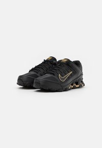 Sapatilhas desportivas pretas com um upper texturizado, detalhes em dourado e uma sola pronunciada projetada para estabilidade e tração. Apresenta atacadores e marcas.