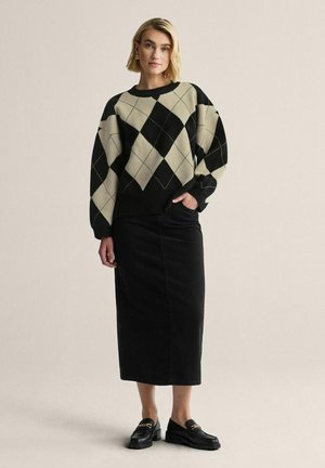Sorte og cremefarvet argyle-mønstret sweater lavet af strikket stof, parret med en sort nederdel. Modellen har sorte loafers på. Ensfarvet baggrund.