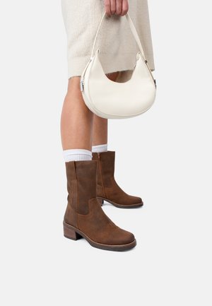 Brown suede ankle boots with a block heel, styled with white socks. Held is a cream-colored crescent-shaped handbag.